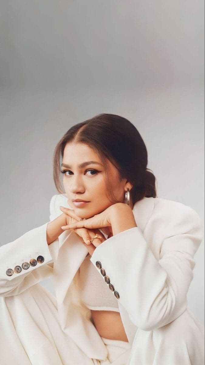 Zendaya posing in an all-white tailored pantsuit leaning chin on hands